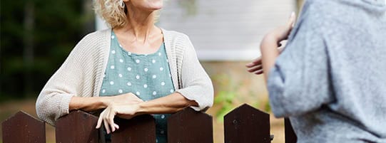 A mature woman with short blonde hair and a light sweater leans on a brown wooden fence, looking off to the side with a slight smile. Her arms are crossed on the fence. Part of a second person is visible on the right, gesturing with their hands as if in conversation with the woman. The background shows trees and a building, suggesting an outdoor, daytime setting.