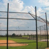 A baseball field with a dirt infield and a manicured grass outfield, surrounded by a tall chain-link fence. The sky above is partly cloudy, and there are light poles in the distance.